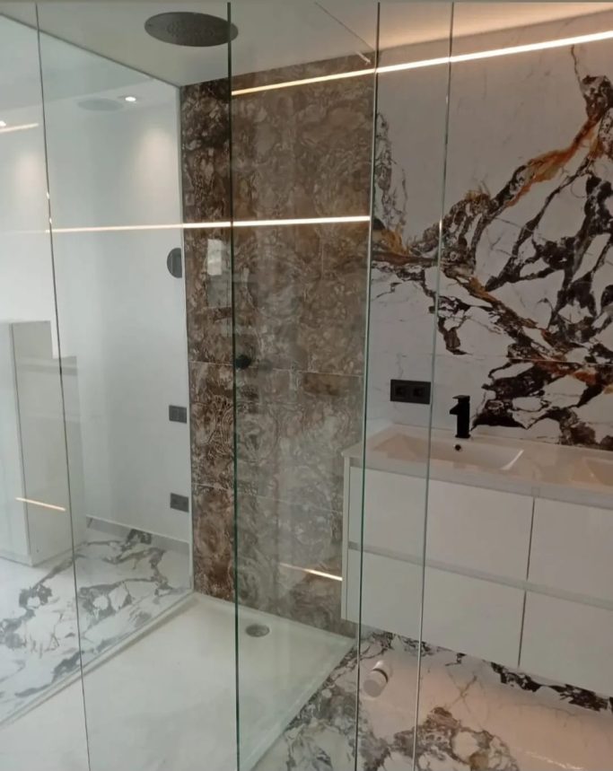 Porcelánicos gran formato Baño moderno con ducha de cristal, paredes de mármol y lavabo blanco.