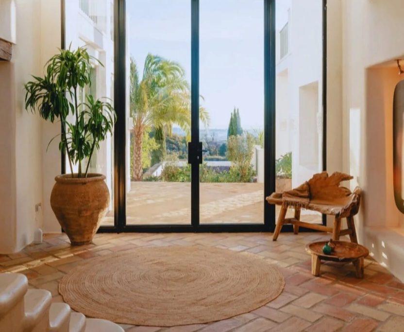 Proyecto de estilo mediterráneo Entrada luminosa con plantas, suelo de barro y grandes ventanales hacia el jardín.